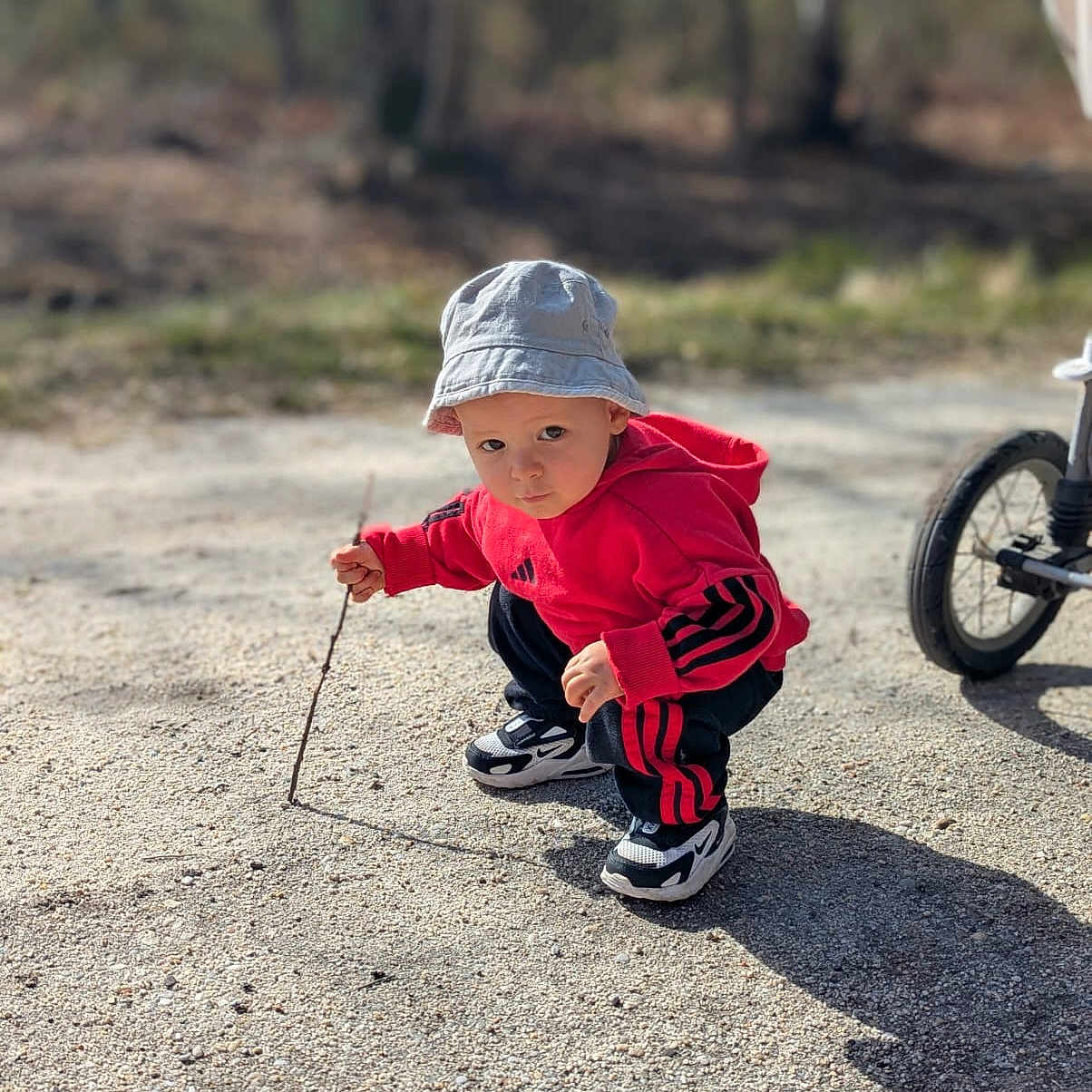 Lino a rejoint le concours — aidez-le/la à gagner de superbes lots ! bicycle_wheel, bucket_hat, candid, child, curious, exploring, gravel_ground, outdoors, park, person, play, portrait, red_hoodie, shadow, sneakers, squatting, stick, sunlight, toddler, trees