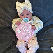 Eliana is registered to the contest to win money with this photo: baby, infant, pacifier, headband, bow, pink, sign, three_months, blanket, sweater, pants, floral_pattern, cute, lying_down, child, adorable, soft_texture, clothing, indoor, portrait