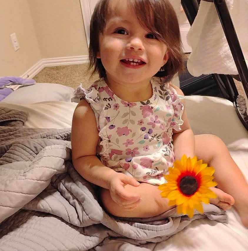 Makenzie is registered to the contest to win money with this photo: toddler, child, smiling, flower, sunflower, bed, blanket, indoor, portrait, happy, clothing, cute, person, hair, face, skin, home, furniture, cozy, casual