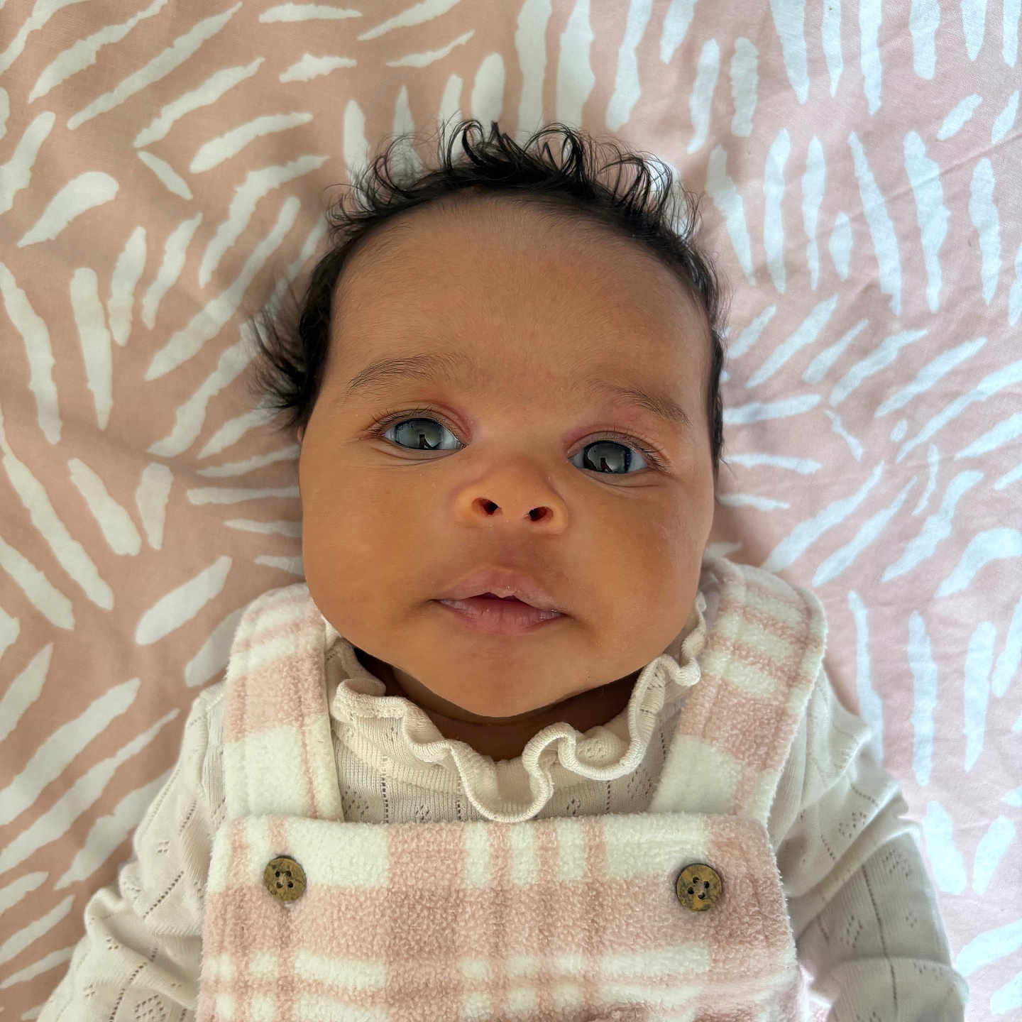 Nala is registered to the contest to win money with this photo: baby, blanket, button, clothing, cute, expression, eyes, face, frilly_collar, hair, infant, lying_down, overalls, pattern, pink, portrait, skin, soft_texture, white, young_child