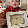 baby, celebration, child, clothing, curtain, expression, floor, gift_box, hands, holiday, home, indoor, packaging, person, plaid_shirt, portrait, present, socks, surprised, young_child