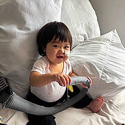 Anna is registered to the contest to win money with this photo: baby, child, infant, smiling, bandage, toy, bed, pillow, white_pillow, sitting, cute, indoors, happy, person, short_hair, black_hair, white_clothing, black_pants, barefoot, cozy