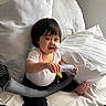 baby, child, infant, smiling, bandage, toy, bed, pillow, white_pillow, sitting, cute, indoors, happy, person, short_hair, black_hair, white_clothing, black_pants, barefoot, cozy