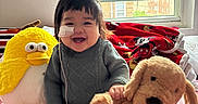 Anna joined the competition — help win amazing prizes! baby, child, smiling, plush_toy, stuffed_animal, yellow_bird, brown_dog, window, blanket, mickey_mouse, bed, indoors, happy, face, bandage, nasal_tube, cute, person, cozy, soft_toys