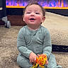 baby, child, onesie, toy, carpet, fireplace, indoor, smiling, sitting, warm, cozy, furniture, floor, happy, young_child, person, playful, electric_fireplace, home, infant