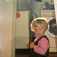 Sindy a rejoint le concours — aidez-le/la à gagner de superbes lots ! child, blonde_hair, pink_shirt, overalls, indoor, washing_machine, cabinet, young_child, curly_hair, appliance, home, person, portrait, floor, furniture, clothing, looking_down, curious, baby, toddler
