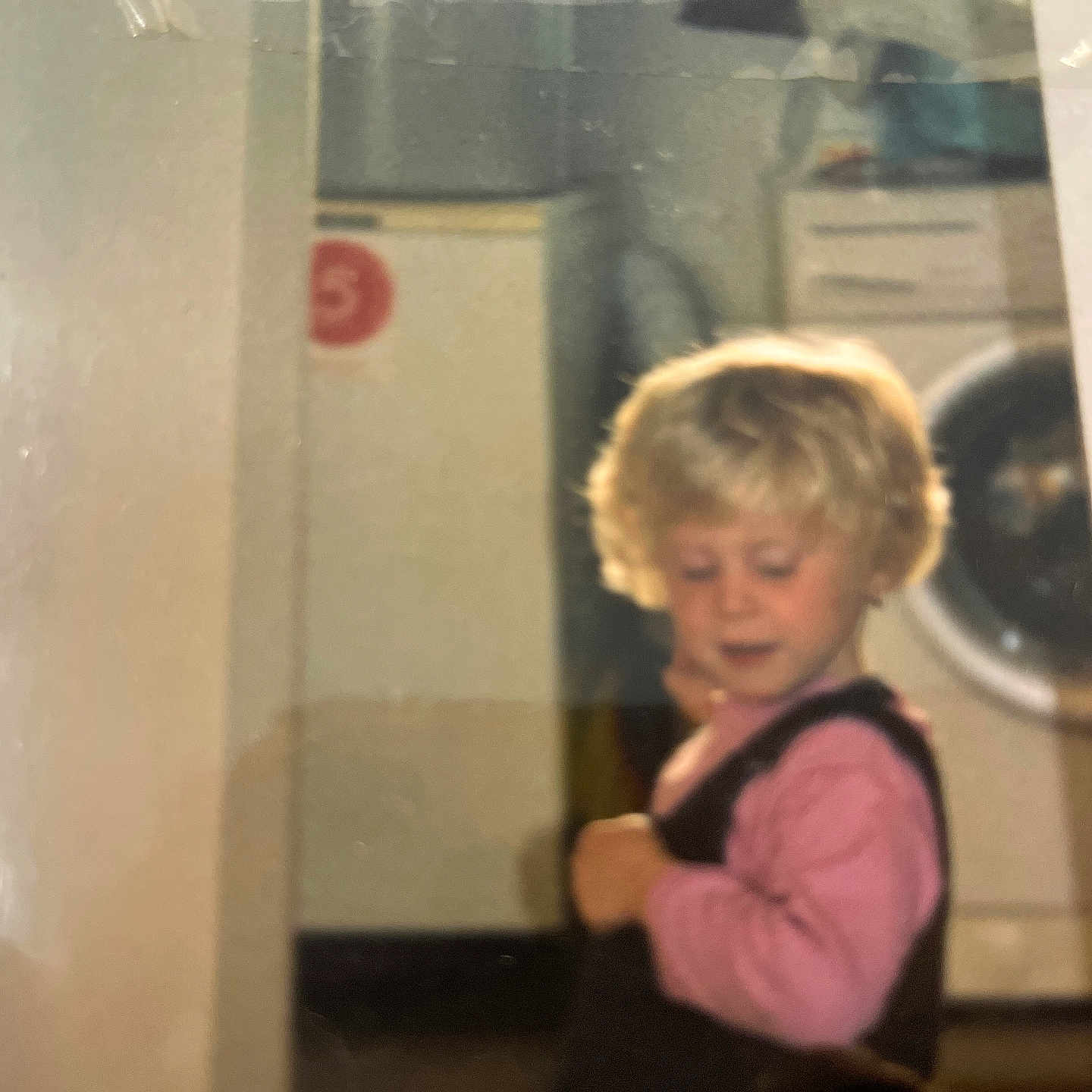 Sindy a rejoint le concours — aidez-le/la à gagner de superbes lots ! appliance, baby, blonde_hair, cabinet, child, clothing, curious, curly_hair, floor, furniture, home, indoor, looking_down, overalls, person, pink_shirt, portrait, toddler, washing_machine, young_child