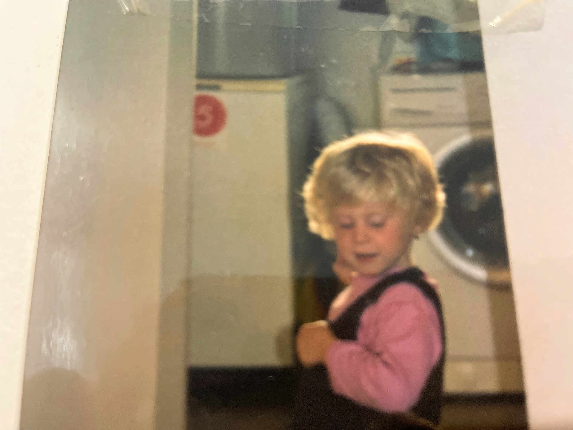Sindy a rejoint le concours — aidez-le/la à gagner de superbes lots ! child, blonde_hair, pink_shirt, overalls, indoor, washing_machine, cabinet, young_child, curly_hair, appliance, home, person, portrait, floor, furniture, clothing, looking_down, curious, baby, toddler