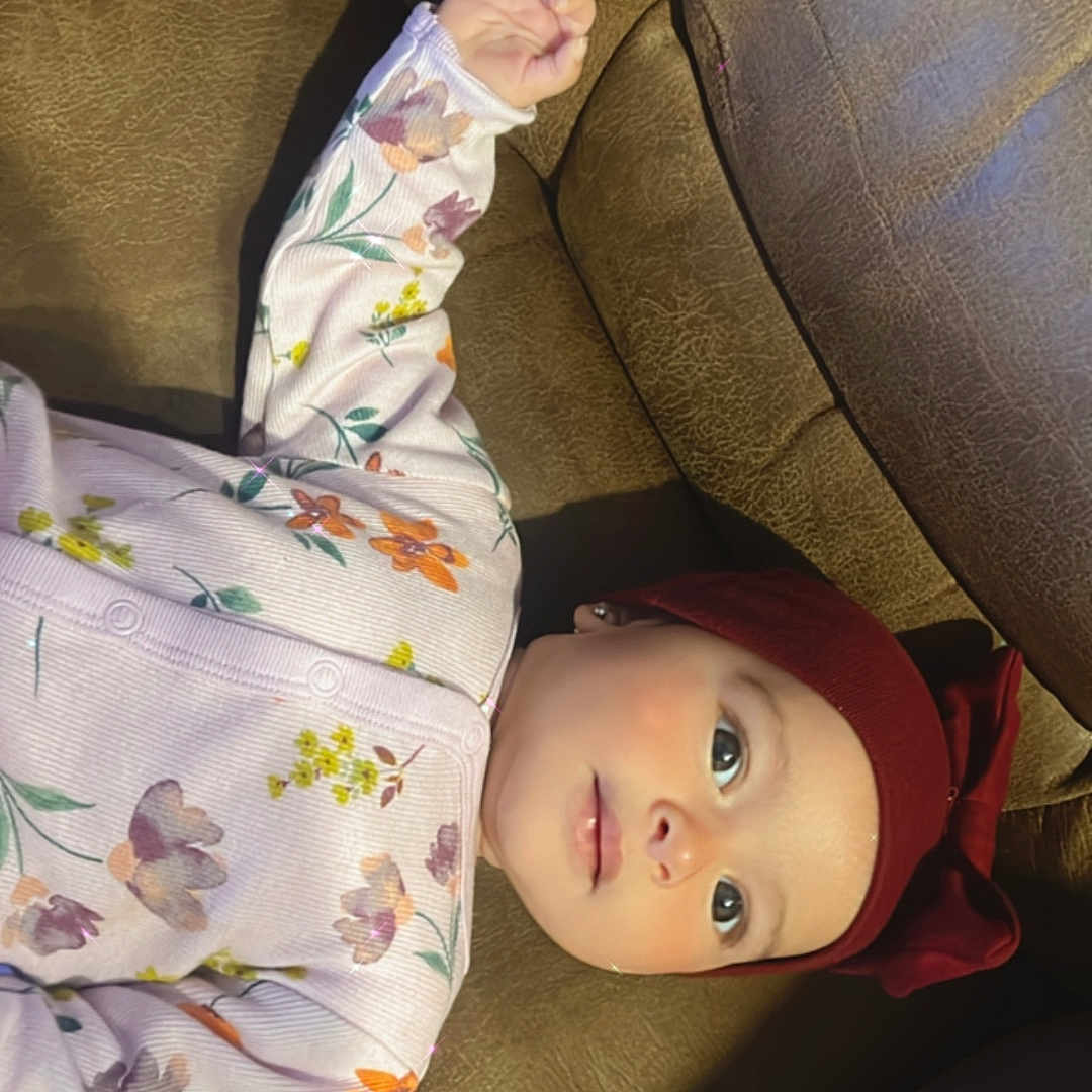 Rana is registered to the contest to win money with this photo: baby, bow, brown_leather, child, comfort, couch, cute, eyes, face, floral_onesie, gazing, headband, indoors, infant, maroon, portrait, skin, sleepwear, small_hand, soft_lighting