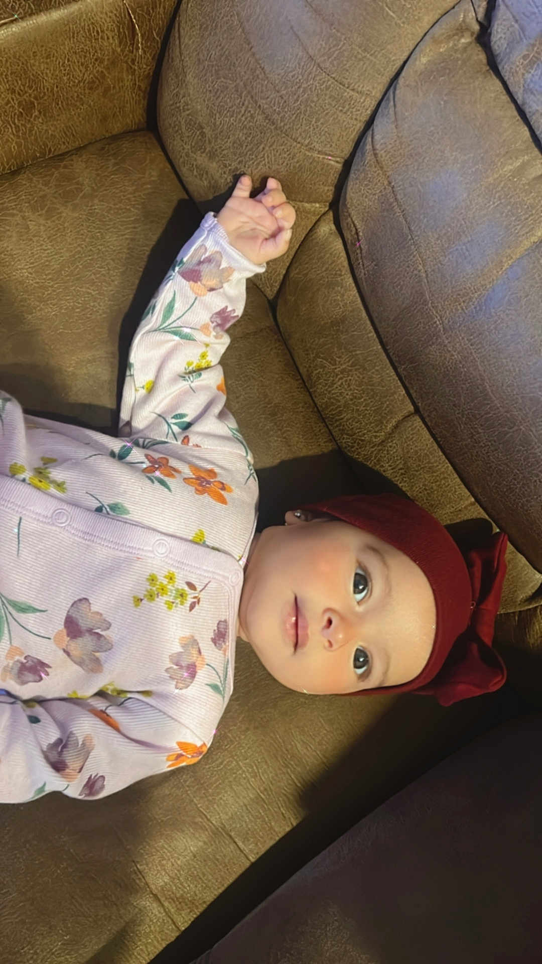 Rana is registered to the contest to win money with this photo: baby, infant, child, couch, brown_leather, floral_onesie, headband, maroon, bow, sleepwear, indoors, portrait, cute, small_hand, gazing, comfort, soft_lighting, skin, face, eyes