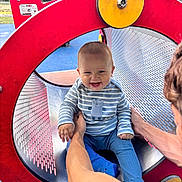 Tommy joined the competition — help win amazing prizes! baby, child, playground, slide, red, blue, striped_clothing, smiling, happy, person, adult_hand, outdoor, metal, play_equipment, park, fun, circular_tunnel, cute, holding, sitting