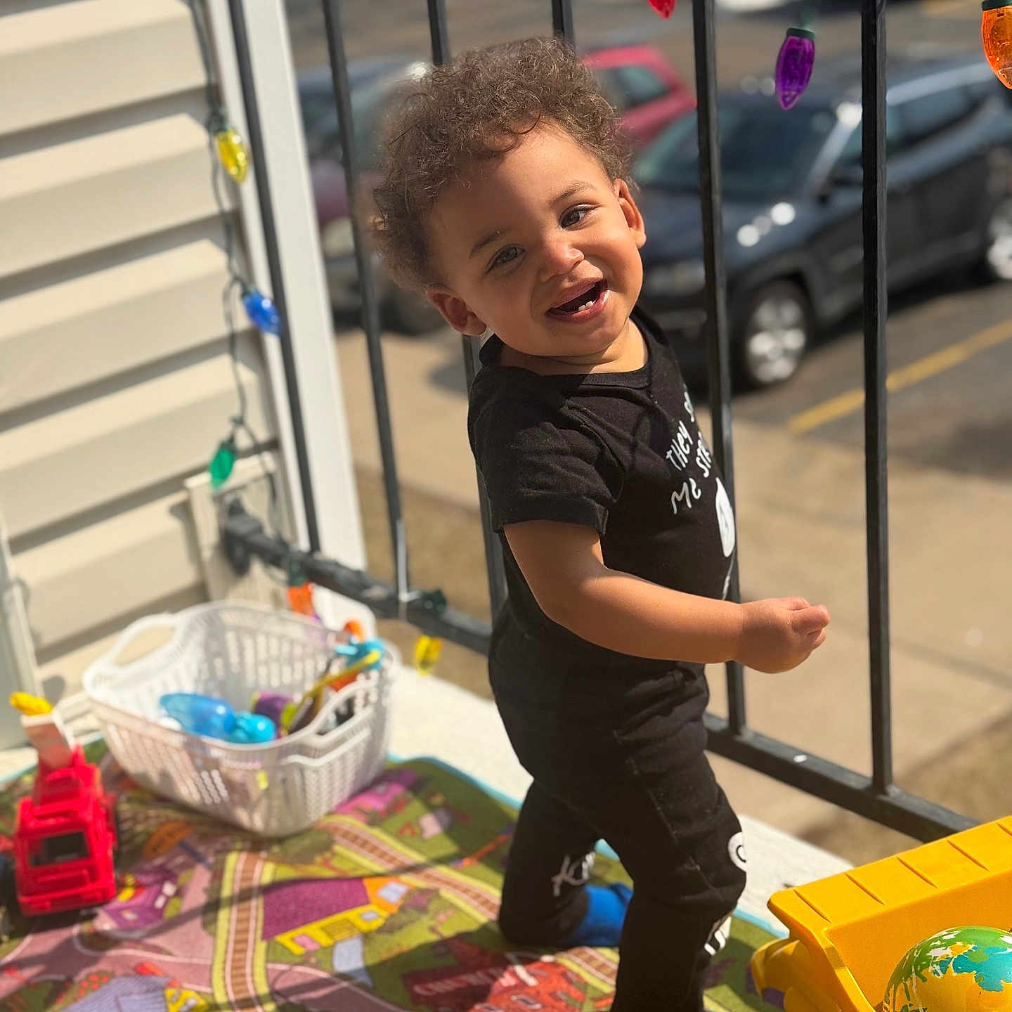 Kimoni joined the competition — help win amazing prizes! toddler, child, curly_hair, standing, play_mat, toys, yellow_truck, basket, colorful_lights, balcony, sunlight, smiling, black_clothing, blue_socks, urban, cars, street, daytime, happy, outdoor