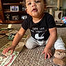 toddler, child, crawling, carpet, book, reading, indoor, living_room, black_shirt, slippers, bear_slippers, curly_hair, face, expression, toy, cat_tree, furniture, floor, person, home