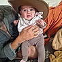 baby, child, hat, cowboy_hat, tattoo, hand, adult, clothing, blanket, expression, surprised, indoor, person, face, legs, sitting, fabric, cozy, holding, portrait