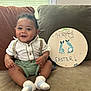 baby, celebration, child, couch, cute, decor, easter_sign, footprints, green_shorts, happy, home, indoor, infant, portrait, seated, shirt, smiling, socks, suspenders, window_blinds