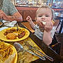 baby_food, casual_clothing, child, cute, dining, eating, food, fork, furniture, high_chair, interior, noodles, person, plate, reflection, restaurant, spoon, table, tattoo, toddler