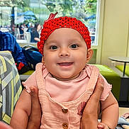 Rishvi is registered to the contest to win money with this photo: baby, child, smiling, red_hat, pink_outfit, hands, indoor, cafe, window, daylight, person, happy, cute, seated, table, people, background, face, portrait, clothing