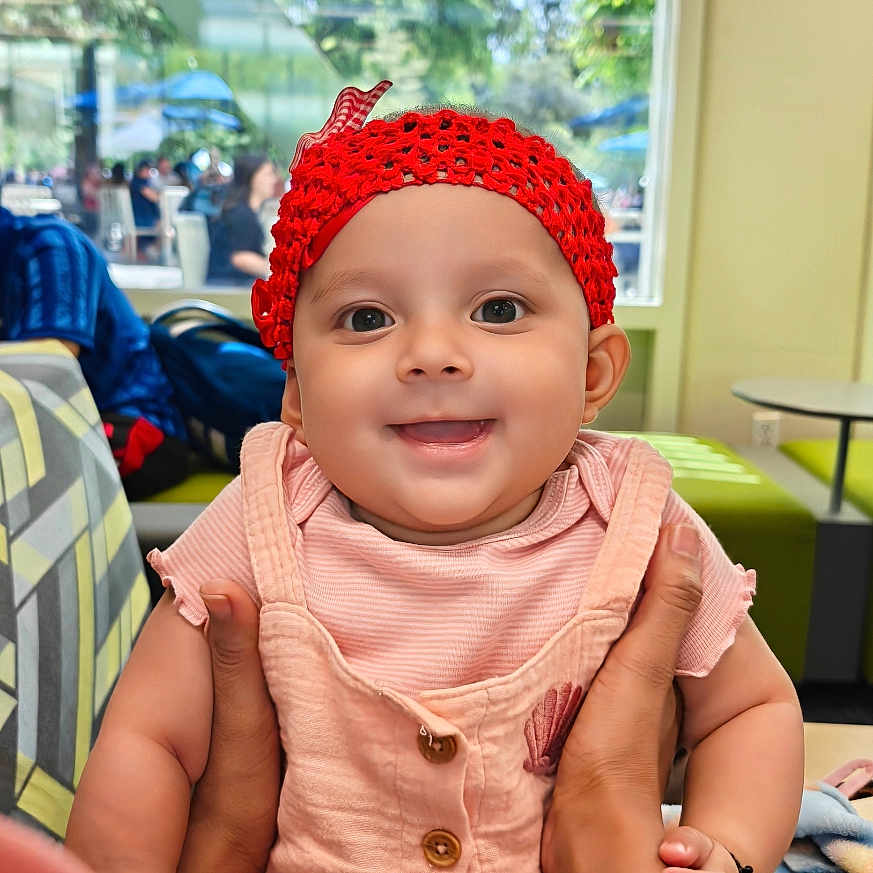 Rishvi is registered to the contest to win money with this photo: baby, child, smiling, red_hat, pink_outfit, hands, indoor, cafe, window, daylight, person, happy, cute, seated, table, people, background, face, portrait, clothing