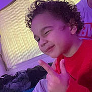 Jordan joined the competition — help win amazing prizes! adult, bed, blanket, casual_clothing, child, cozy, curly_hair, curtain, face, hand_gesture, home, indoor, peace_sign, person, portrait, purple_light, red_sweater, relaxed, smiling, winking