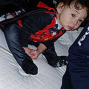 Legend joined the competition — help win amazing prizes! toddler, child, costume, red, black, mattress, bedroom, indoor, curly_hair, socks, pouting, person, crouching, clothing, fabric, furniture, soft_lighting, casual, home, curious