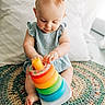 baby, child, headband, bow, blue_dress, toy, stacking_toy, colorful, sitting, rug, floor, indoor, pillow, curious, playing, infant, hands, feet, concentration, cute