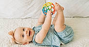 Margaret joined the competition — help win amazing prizes! baby, ball, blue_clothing, child, closeup, cute, face, feet, hands, happy, headband, indoor, infant, lying_down, person, playful, quilted_bedspread, skin, smiling, toy