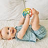 baby, infant, child, toy, ball, headband, blue_clothing, smiling, lying_down, quilted_bedspread, cute, happy, playful, indoor, closeup, person, skin, face, hands, feet