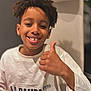 boy, casual_clothing, chair, child, closeup, curly_hair, expression, happy, head_and_shoulders, indoor, person, portrait, positive_gesture, seated, smile, text_on_clothing, thumbs_up, warm_lighting, white_sweatshirt, young