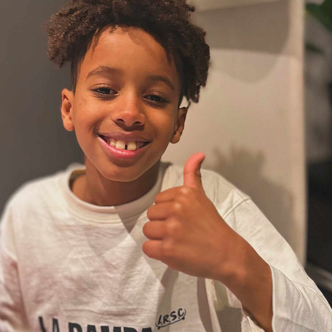 Denilson participe au concours pour gagner de l'argent avec cette photo : boy, casual_clothing, chair, child, closeup, curly_hair, expression, happy, head_and_shoulders, indoor, person, portrait, positive_gesture, seated, smile, text_on_clothing, thumbs_up, warm_lighting, white_sweatshirt, young
