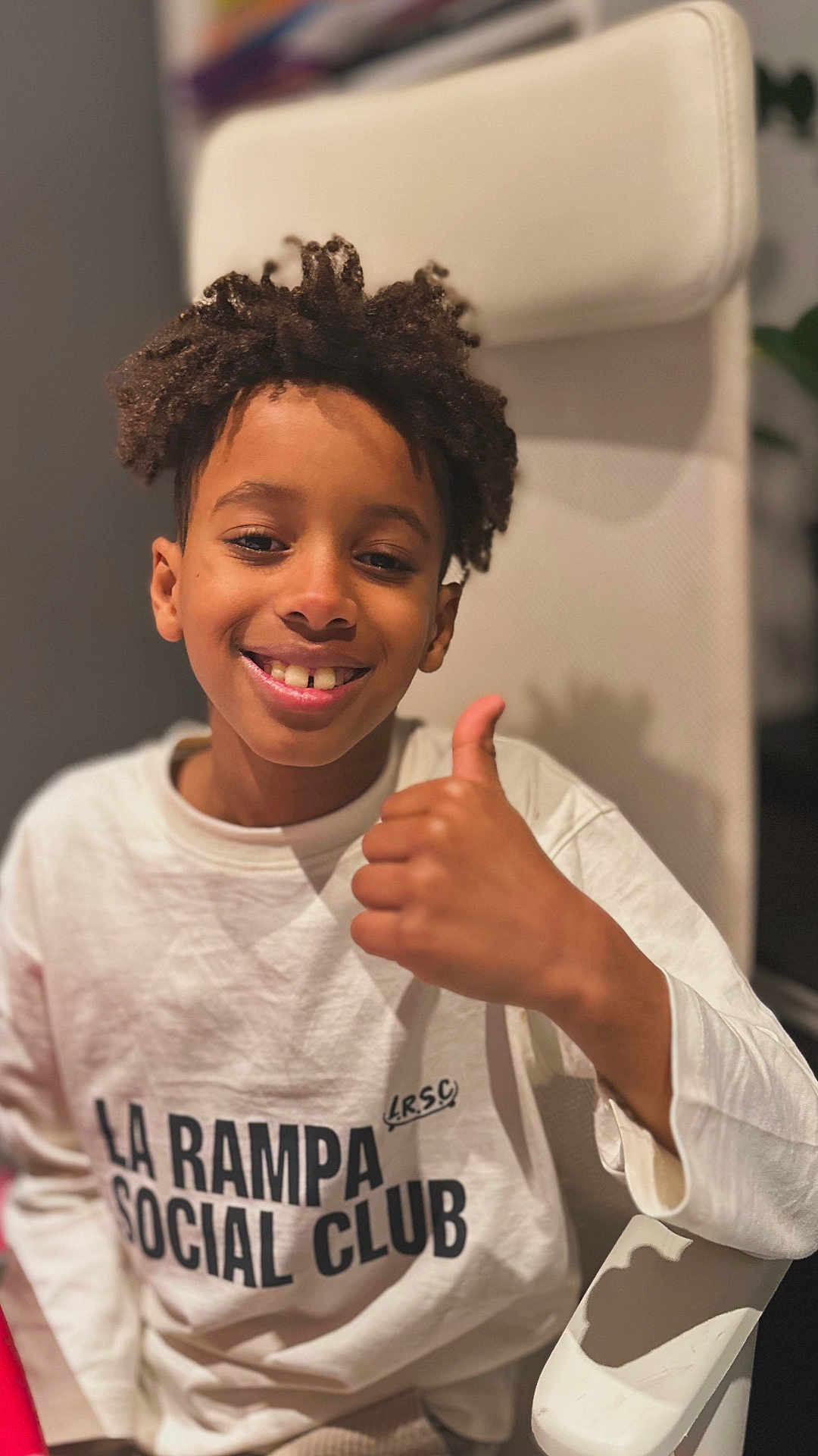 Denilson participe au concours pour gagner de l'argent avec cette photo : boy, casual_clothing, chair, child, closeup, curly_hair, expression, happy, head_and_shoulders, indoor, person, portrait, positive_gesture, seated, smile, text_on_clothing, thumbs_up, warm_lighting, white_sweatshirt, young