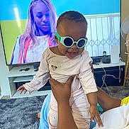 Arbela is registered to the contest to win money with this photo: baby, sunglasses, adult_hand, television, woman, carpet, room, home, furniture, smile, clothing, person, technology, indoor, playful, holding, cord, remote_control, couch, happy