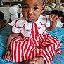 Arbela is registered to the contest to win money with this photo: baby, child, indoor, dress, red_stripes, white_collar, sitting, blue_quilt, foot, hand, expression, portrait, curious, face, clothing, person, home, furniture, room, soft_lighting