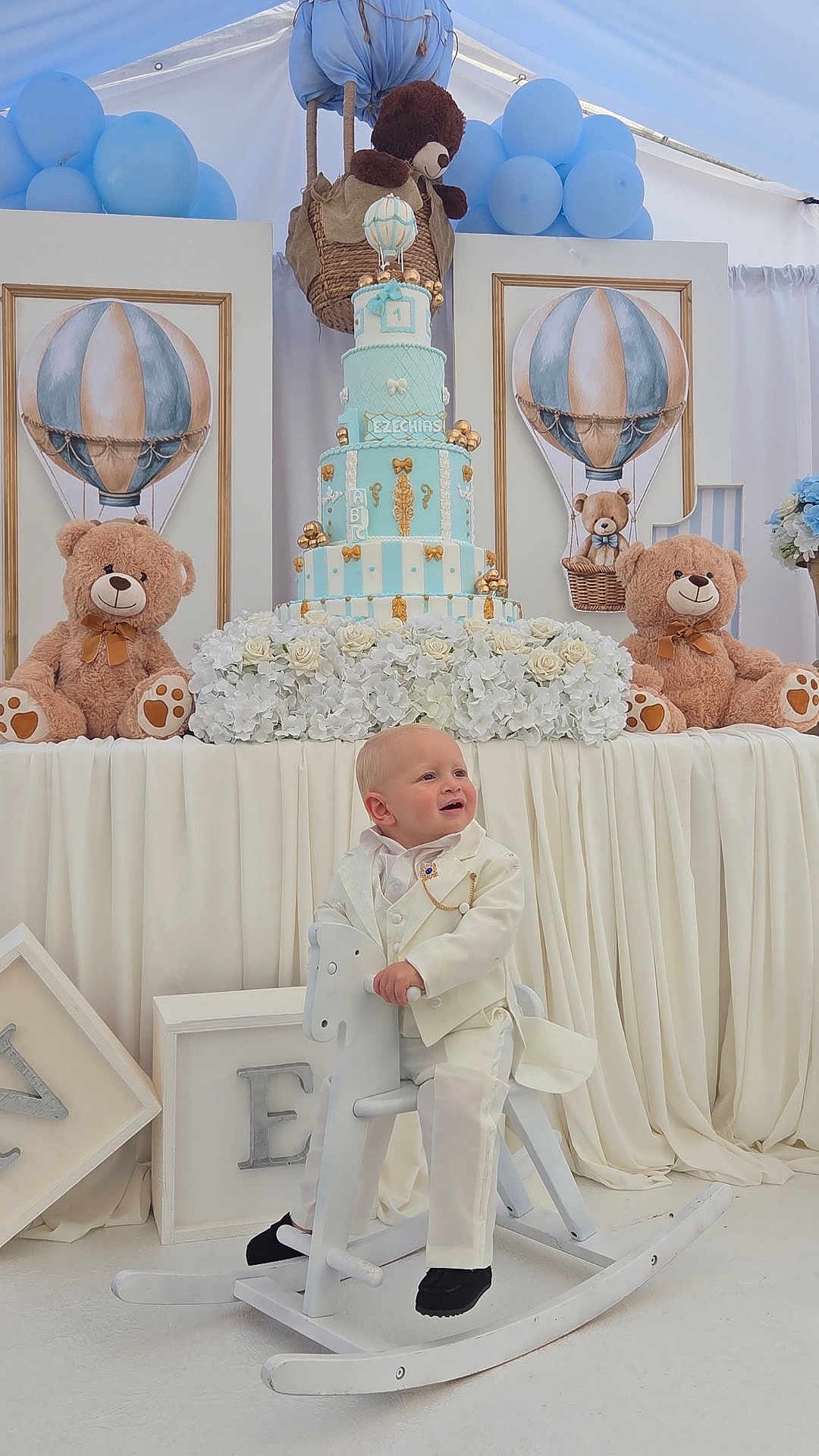 Ezéchias a rejoint le concours — aidez-le/la à gagner de superbes lots ! toddler, child, white_suit, rocking_horse, birthday_cake, teddy_bear, balloons, flowers, decorations, party, celebration, indoor, smiling, happy, white_background, wooden_toy, formal_wear, blue, cream, festive