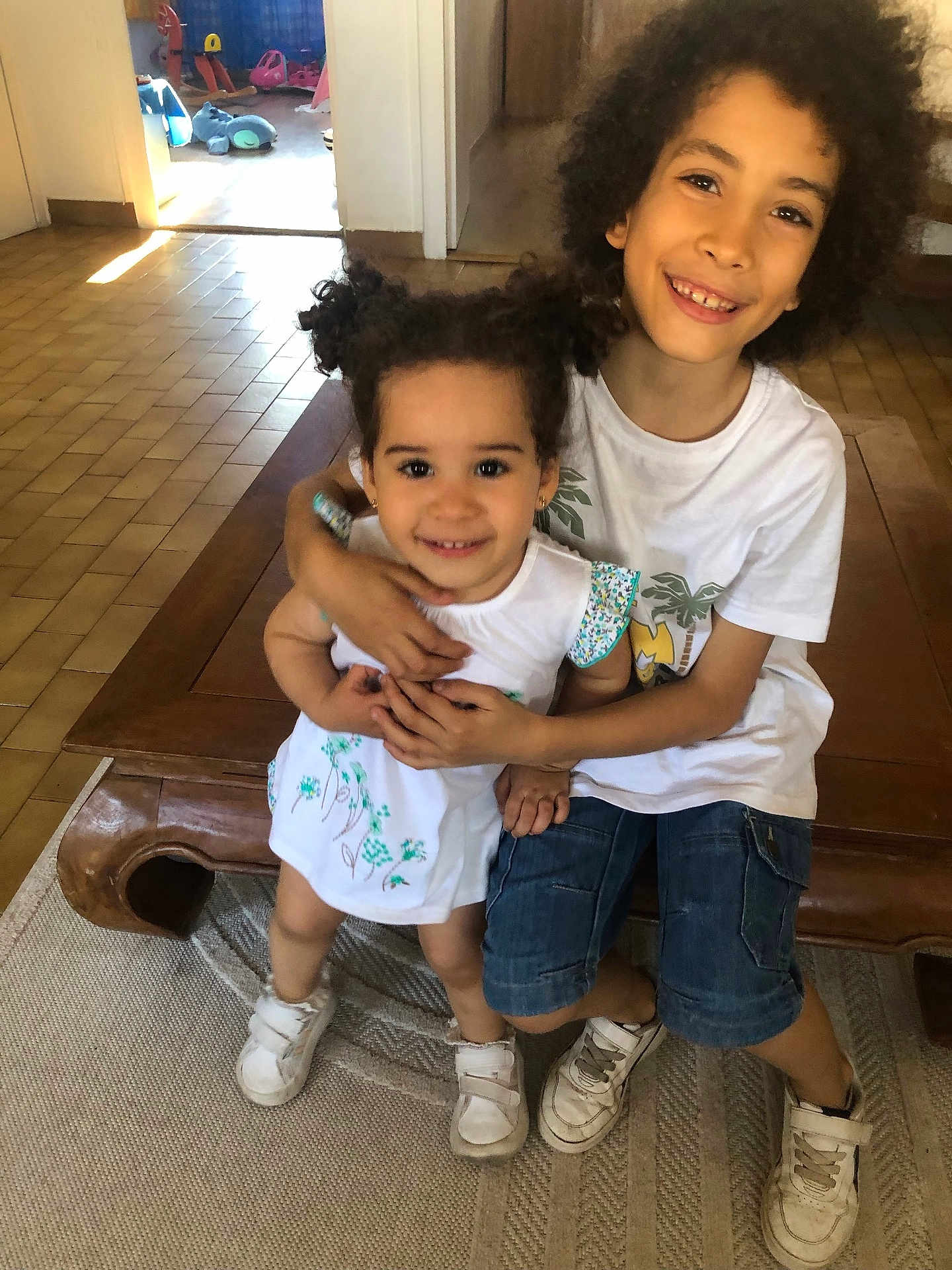 Yara Carneiro participe au concours pour gagner de l'argent avec cette photo : child, children, siblings, hug, smile, indoor, wooden_bench, curly_hair, pigtails, white_shirt, denim_shorts, sneakers, playroom, toys, floor_tiles, doorway, carpet, happy, family, portrait