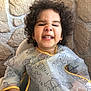 child, toddler, curly_hair, smiling, high_chair, bib, bear_pattern, face, teeth, earrings, hand, stone_wall, indoor, cute, happy, playful, portrait, clothing, baby, person