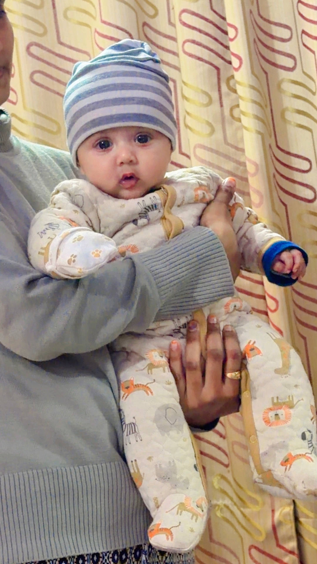 Atharv is registered to the contest to win money with this photo: adult, animal_print, baby, child, cozy_clothing, curtain, cute, expression, face, hand, hat, holding, indoor, infant, onesie, person, portrait, striped_hat, sweater, warm_lighting