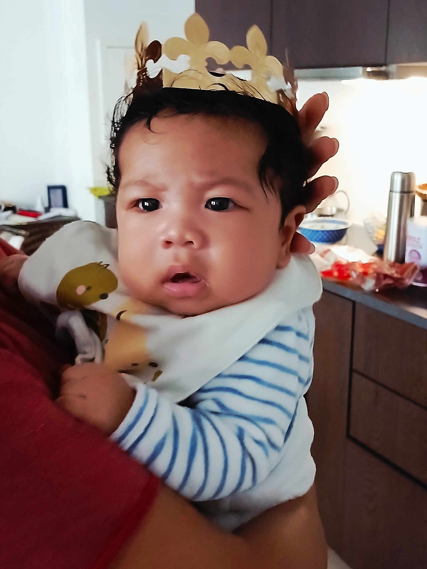 Gael participe au concours pour gagner de l'argent avec cette photo : baby, child, crown, striped_clothing, bib, indoor, kitchen, person, holding, face, curious, expression, hand, adult, furniture, cabinet, bowl, thermos, food, home