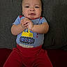 baby, child, infant, clapping, smiling, blue_shirt, red_pants, excavator, sitting, couch, indoor, face, hands, happy, young, person, casual_clothing, home, portrait, cute