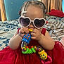 Azariah joined the competition — help win amazing prizes! toddler, child, toy, heart_shaped_sunglasses, red_dress, braided_hair, indoors, bed, blanket, colorful, playful, cute, person, infant, baby, fashion, accessory, smile, fun, home