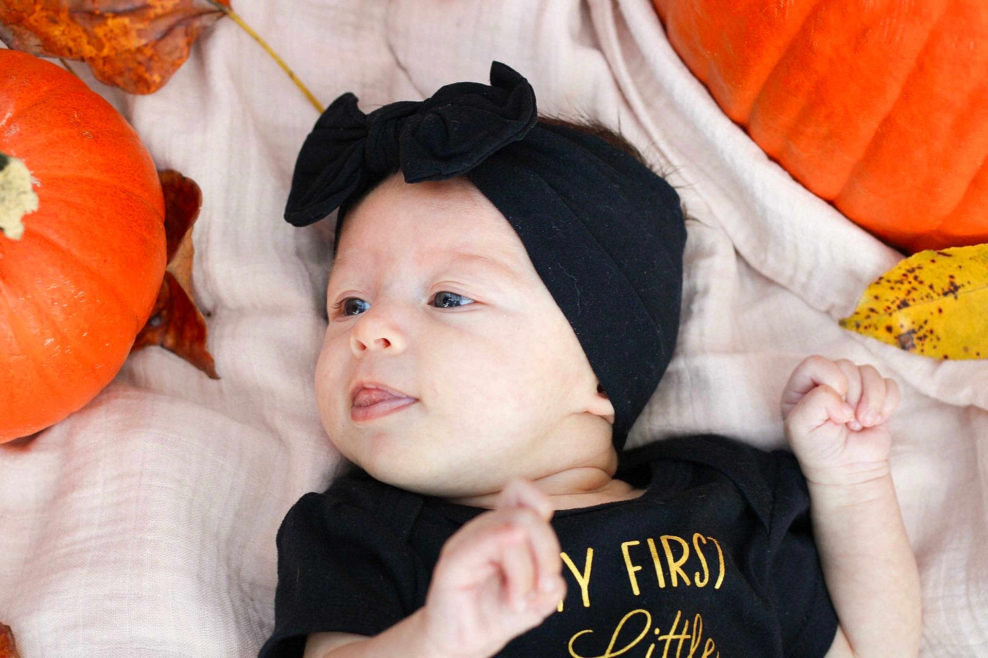 Lillianna is registered to the contest to win money with this photo: baby, infant, headband, bow, black_clothing, pumpkin, autumn_leaves, blanket, lying_down, tongue_out, cute, playful, fall, seasonal, child, portrait, soft_texture, orange, black, skin