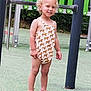 child, toddler, smiling, playground, romper, giraffe_pattern, pink_sandals, curly_hair, outdoor, green_background, metal_play_structure, happy, standing, summer_clothing, person, feet, legs, nature, daylight, recreation
