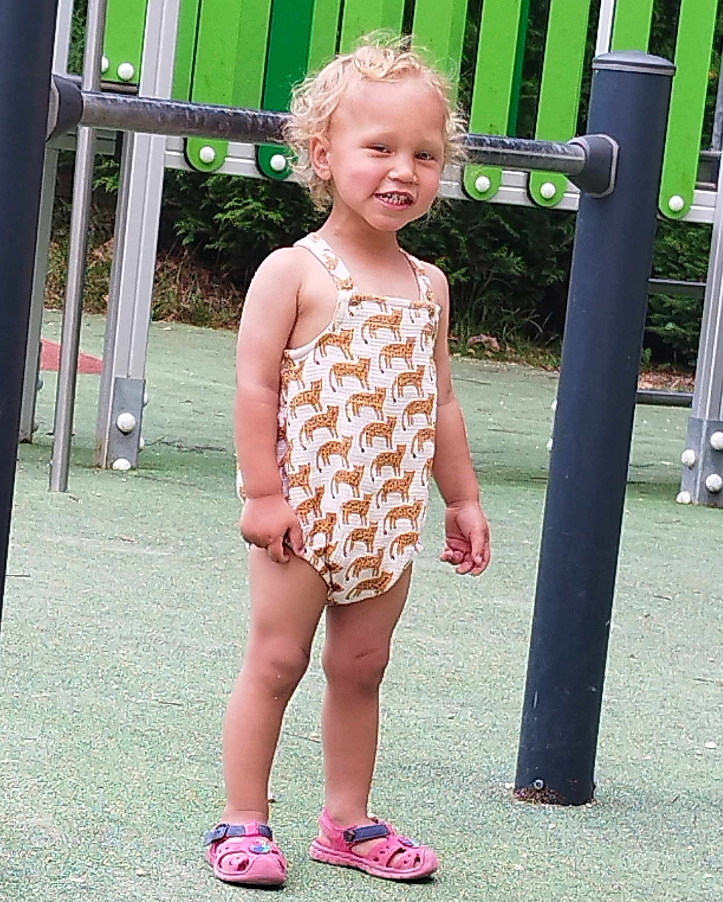 Yaël a rejoint le concours — aidez-le/la à gagner de superbes lots ! child, toddler, smiling, playground, romper, giraffe_pattern, pink_sandals, curly_hair, outdoor, green_background, metal_play_structure, happy, standing, summer_clothing, person, feet, legs, nature, daylight, recreation