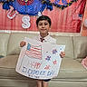 child, boy, independence_day, american_flag, poster, celebration, holiday, festive, decorations, sofa, home_interior, carpet, smiling, shorts, shirt, tie, fireworks, patriotic, handmade_sign, happy