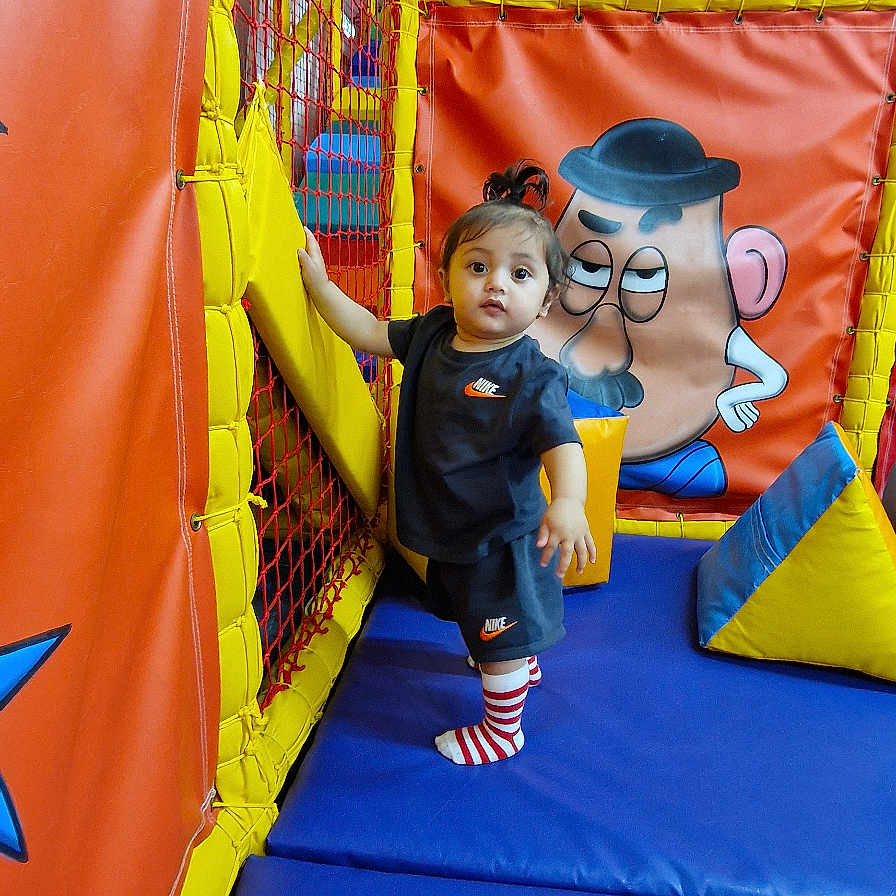 Yazen is registered to the contest to win money with this photo: toddler, child, indoor_play_area, playground, netting, colorful, padded_walls, cartoon_art, blue_floor, red_padding, yellow_padding, striped_socks, sporty_clothing, curious_expression, standing, hand_grip, play_area, soft_play, fun, young_child