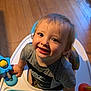child, toddler, smiling, baby_walker, toy, indoor, wooden_floor, curly_hair, gray_shirt, nike_logo, playful, happy, cute, person, young_child, close_up, looking_up, bright, colorful, face