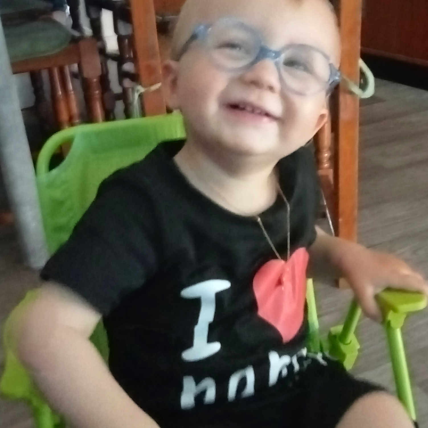Timéo a rejoint le concours — aidez-le/la à gagner de superbes lots ! black_shirt, casual_clothing, chair, child, floor, furniture, glasses, green_chair, happy, heart_graphic, home, indoor, person, portrait, seated, shorts, smile, toddler, wooden_chair, young_child