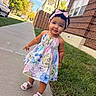 toddler, child, sidewalk, dress, floral_pattern, headband, pink_bow, sandals, grass, brick_building, tree, sunlight, outdoor, curly_hair, smile, cute, urban, daytime, person, portrait