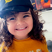 Ilyana a rejoint le concours — aidez-le/la à gagner de superbes lots ! child, toddler, smiling, curly_hair, cap, hat, yellow_shirt, indoor, happy, face, cute, person, clothing, portrait, young_child, expression, joyful, childhood, casual, close_up