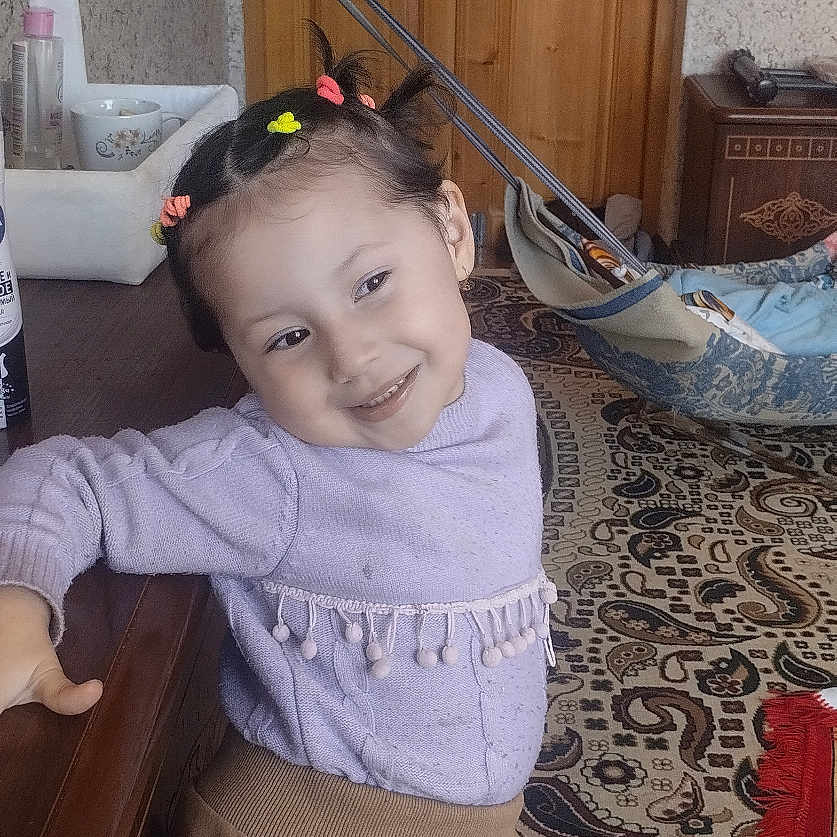 Mubina is registered to the contest to win money with this photo: carpet, child, cute, furniture, hair_ties, happy, home, indoor, leaning, pants, patterned_rug, person, playful, purple_chair, room, smiling, socks, sweater, wooden_doors, wooden_dresser