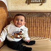 Tiago a rejoint le concours — aidez-le/la à gagner de superbes lots ! baby, child, smile, hoodie, bear_graphic, wicker_sofa, cushion, indoor, signs, wooden_signs, text, cozy, happy, seated, cute, portrait, infant, clothing, face, person